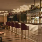 The Carlton Milan hotel bar featuring a backlit bottle display, elegant bar stools, sculptural ceiling lights, and contemporary furnishings