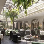 Bright indoor lounge at The Carlton Milan beneath a glass roof, with leafy plants, arched windows, and comfortable seating