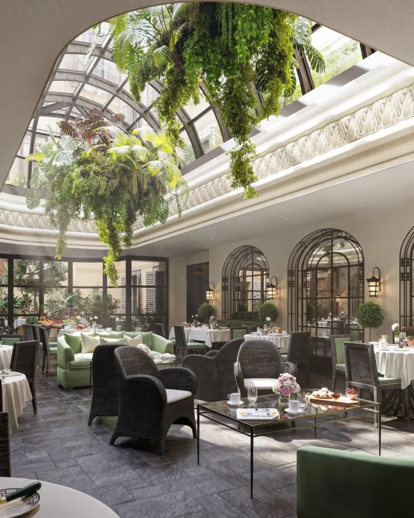 Bright indoor lounge at The Carlton Milan beneath a glass roof, with leafy plants, arched windows, and comfortable seating