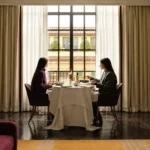 Elegant restaurant table beside a large sunlit window at The Carlton Milan, surrounded by curtains and sophisticated furnishings
