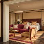 Warm luxury suite interior at The Carlton Milan featuring a king-size bed, burgundy sofa, patterned rug, and elegant contemporary furnishings